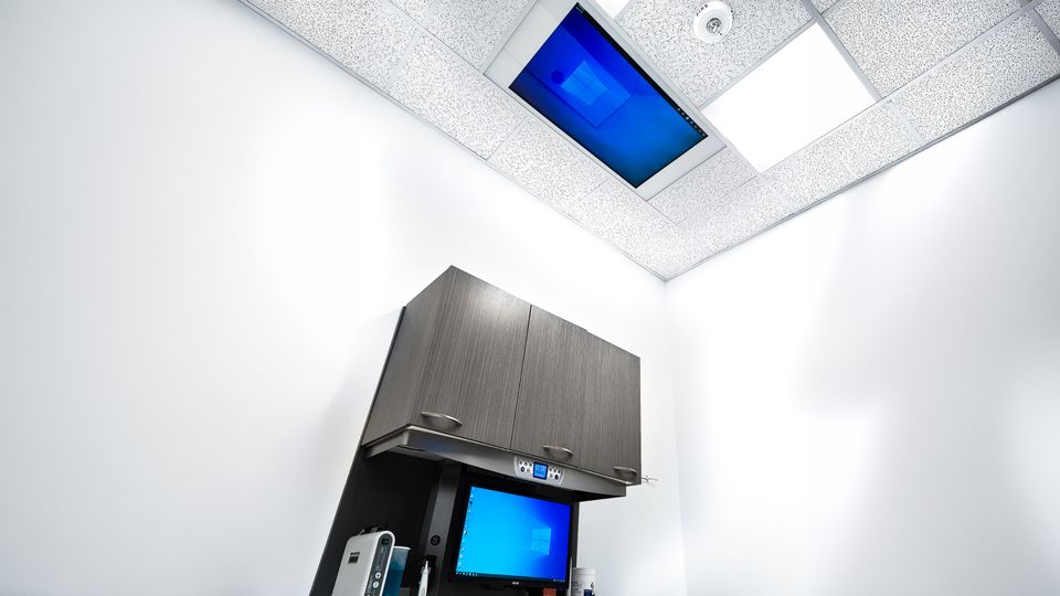 Dental treatment room with ceiling-mounted TV screen and wall cabinets above workstation